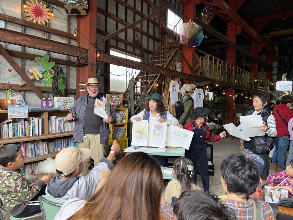 床に座る子どもたちの前で、マイクを持って絵本を掲げる読み聞かせの人。