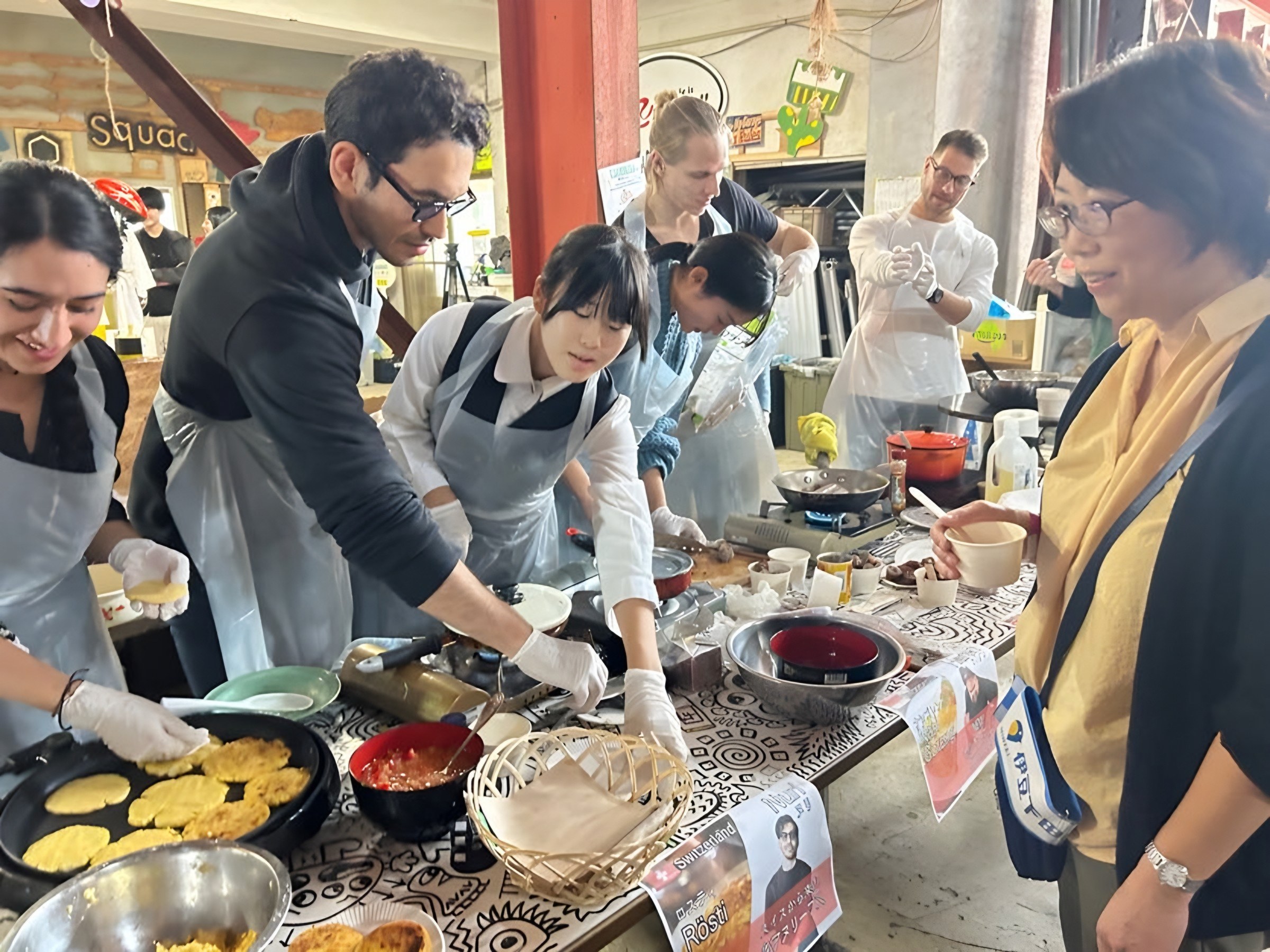 International participants and local residents cooking side by side at a long table of shared dishes.